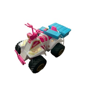 Könnte beinhalten: Ein weißes, pinkes und blaues Spielzeug-Geländefahrzeug mit dem Barbie-Logo auf der Vorderseite. Das Fahrzeug hat große schwarze Reifen mit goldenen Felgen, eine blaue Aufbewahrungsbox auf der Rückseite und eine weiße Schnur um Lenker und Sitz.