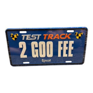 Puede incluir: Una placa de matrícula rectangular azul con el texto TEST TRACK en plata y naranja, el texto 2 GOO FEE en blanco y la palabra Epcot en letras blancas pequeñas. Dos iconos de Mickey Mouse a cuadros negros y amarillos están en las esquinas.