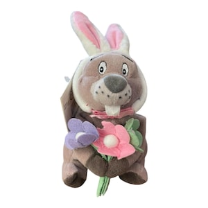 Könnte beinhalten: Ein Plüschhase mit hellbraunem Körper, rosa gefütterten Ohren und einer grauen Nase. Er hält einen Strauß lila und rosa Blumen mit grünen Stielen. Der Hase trägt eine rosa Fliege.
