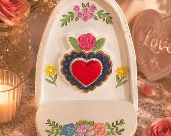Può includere: Una mensola da parete ad arco bianca con un emblema centrale a forma di cuore rosso in rilievo, circondato da un bordo smerlato blu scuro. La mensola è decorata con motivi floreali dipinti in rosa, giallo, blu e verde, inclusa una rosa.