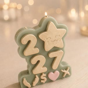 May include: A sage green candle shaped like a cloud with a lit wick. It features gold-coloured raised numbers 2, 2, and 7, a gold star with a smiling face, and small gold symbols including hands, a dollar sign, a heart, and an aeroplane.