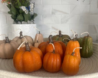 May include: A collection of small, hand-crafted pumpkin and gourd-shaped candles in various shades of orange, green, and brown. The candles are arranged in a white woven bowl.