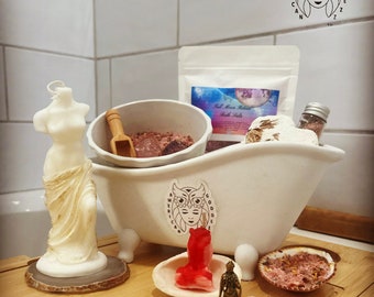 Peut inclure: Une baignoire blanche remplie de sels de bain roses, une petite pelle en bois et un sac blanc de sels de bain étiqueté "Full Moon Ritual Bath Salts". La baignoire est sur une surface en bois avec une statue blanche, une bougie rouge, une petite statue dorée et un rocher blanc.