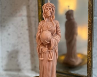 Può includere: Una candela rosa a forma di donna che tiene una bilancia e un globo. La candela è su un piatto dorato e davanti a uno specchio.