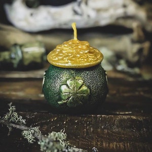 May include: A green and gold candle shaped like a pot of gold with a four-leaf clover design. The candle has a gold lid with a textured design.