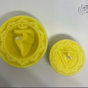 May include: Two yellow candles with a raised design of a chakra symbol and a wick in the center. The larger candle has a more detailed design.