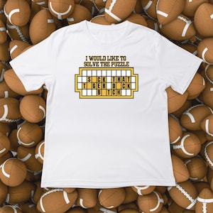 May include: White t-shirt with a humorous graphic. The graphic features a puzzle-like design with the text "I WOULD LIKE TO SOLVE THE PUZZLE" above a grid that spells out "SICK THAT TIGER DICK BITCH".