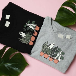 May include: Two t-shirts, one black and one gray, each featuring a graphic of a ghost among potted plants. The design includes various green plants and terracotta pots, creating a whimsical and spooky theme. The shirts are folded on a pink surface.