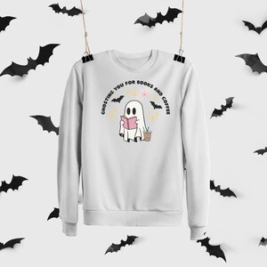 Könnte beinhalten: Weißes Sweatshirt mit einem Geister-Motiv, das ein rosa Buch liest und einen Kaffeebecher hält. Der Text lautet "Ghosting you for books and coffee". Schwarze Fledermäuse sind um das Sweatshirt herum verteilt.