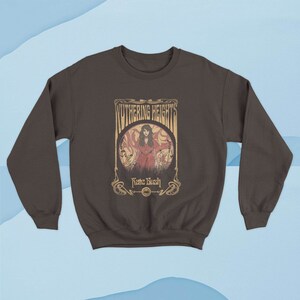 May include: A brown sweatshirt with a vintage-style graphic of a woman in a red dress standing in a field. The graphic includes the text "Wuthering Heights" and "Kate Bush".