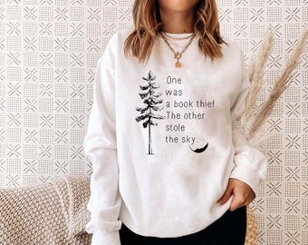 Puede incluir: Sudadera blanca con un gráfico de árbol negro y la cita: "One was a book thief. The other stole the sky." La sudadera es de cuello redondo y tiene mangas largas. El fondo es una pared estampada.