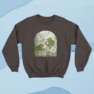May include: A brown sweatshirt with a graphic print of two frogs in a garden setting. The frogs are green and wearing hats. The graphic is surrounded by a white border.