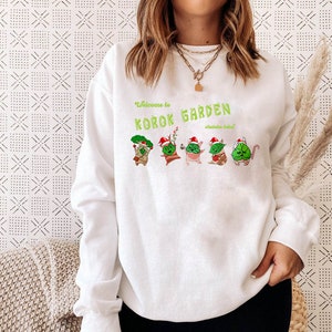 May include: White sweatshirt with a green and red graphic of a Christmas-themed "Korok Garden" with the text "Welcome to Korok Garden - celebrate Zelda!"