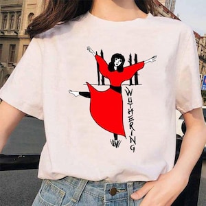 Puede incluir: Camiseta blanca con un gráfico de una mujer con un vestido rojo, leggings negros y la palabra "WUTHERING" en negro. El diseño incluye líneas verticales negras y hierba.