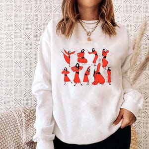 Puede incluir: Sudadera blanca con un gráfico de mujeres con vestidos rojos en varias poses. La ilustración es minimalista, con contornos negros y rojo liso. La sudadera es de estilo casual y cómodo.