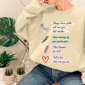 Könnte beinhalten: Ein hellbeiger Pullover mit einem grafischen Druck eines Herzens, eines Rosmarinzweigs, eines Lavendelzweigs und eines Kristalls. Der Text auf dem Pullover lautet: "Wirf immer verschüttetes Salz über deine linke Schulter. Halte Rosmarin an deinem Gartentor. Pflanze Lavendel, für Glück. Verliebe dich, wann immer du kannst."