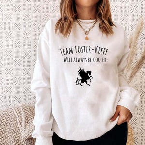 May include: White sweatshirt with the text "Team Foster-Keefe Will Always Be Cooler" and a black winged horse graphic. The long-sleeved top is a casual style.