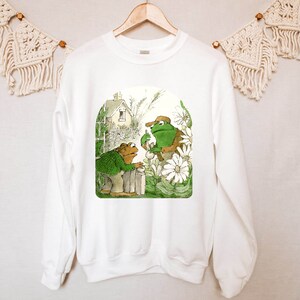 May include: White sweatshirt with a green frog and brown frog illustration. The frogs are standing near a white picket fence with flowers and a house in the background. The text "Frog and Toad are Friends" is printed on the sweatshirt.