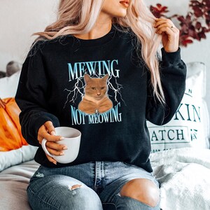 May include: Black sweatshirt featuring the text "MEWING NOT MEOWING" and an orange cat graphic. The text is in light blue with lightning bolt details. A casual style sweatshirt.