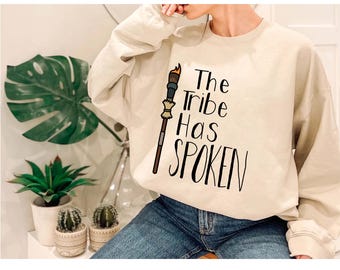 May include: Beige sweatshirt with the text "The Tribe Has Spoken" in black script. A torch graphic is on the left side of the text. The sweatshirt is being worn by a person wearing blue jeans.