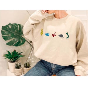 May include: Beige crewneck sweatshirt with colorful graphic icons: a wheat stalk, a blue fish, red berries, a gray mouse, and a green slug. The sweatshirt is worn with blue jeans. Decorative plants are in the background.