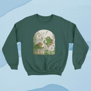 May include: A green sweatshirt with a vintage illustration of two frogs in a garden setting. The frogs are wearing hats and holding a cup of tea. The illustration is surrounded by a white border.