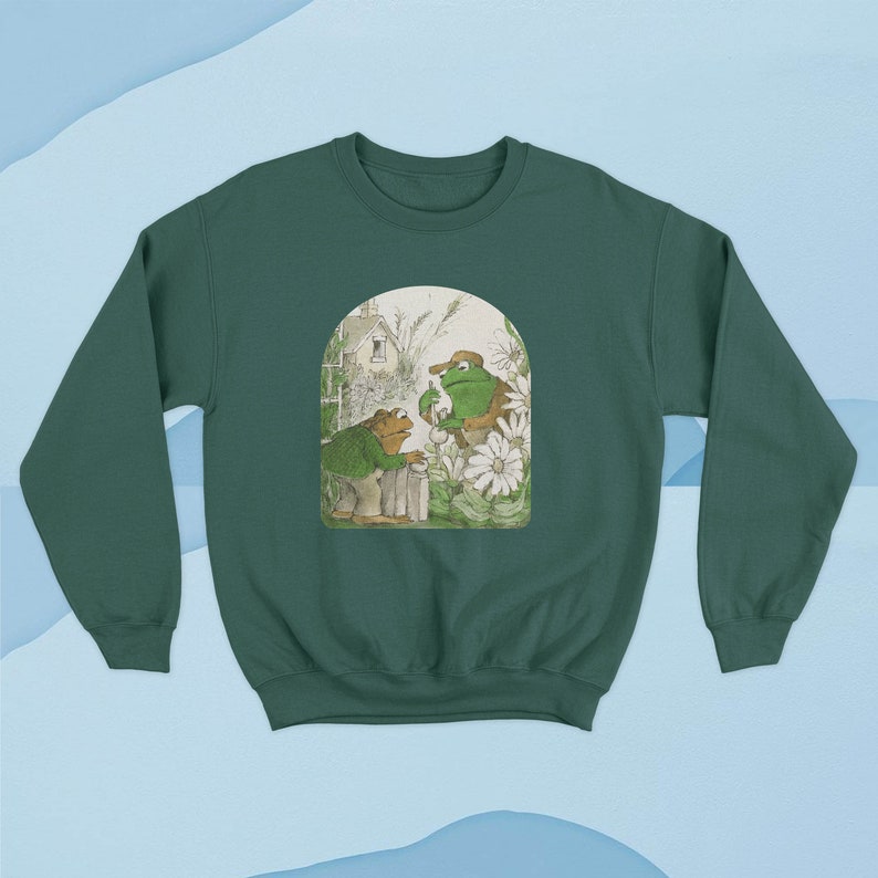May include: A green sweatshirt with a vintage illustration of two frogs in a garden setting. The frogs are wearing hats and holding a cup of tea. The illustration is surrounded by a white border.