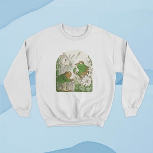 May include: White sweatshirt with a green frog illustration. The frog is wearing a hat and holding a cup. The frog is standing in a garden with flowers and a small house in the background.