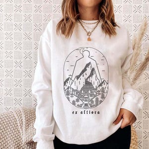 May include: White sweatshirt with a black and white mountain and landscape design. The graphic includes a mountain range, trees, and a building, with the text "ex altiora" below the design. The sweatshirt is long-sleeved.