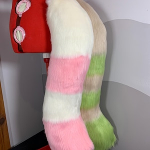 Peut inclure: Deux queues en fausse fourrure. L'une est rayée blanche, rose et blanche. L'autre est rayée beige et verte. Les queues sont exposées sur un mannequin rouge. Le fond est un mur gris et blanc.
