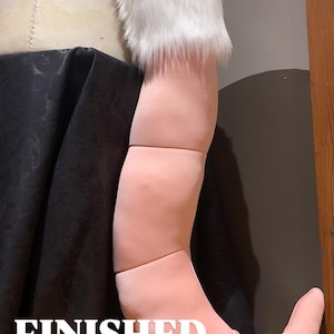 May include: A pink and white faux fur tail with a curved shape. The tail is made of soft, plush fabric and has a realistic look. The tail is attached to a black garment.