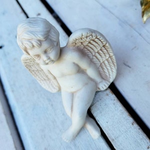 May include: A white ceramic angel figurine with wings and a sad expression. The angel is sitting on a white wooden surface.