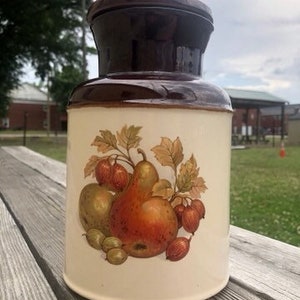 May include: A vintage ceramic cookie jar with a brown lid and a white body. The jar features a colorful illustration of pears, apples, and berries.