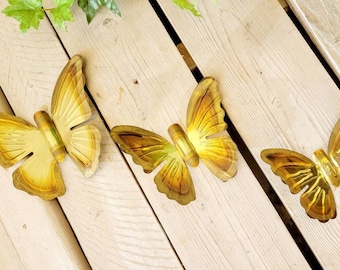 May include: Three gold metal butterflies of different sizes are attached to a wooden surface. The butterflies have a textured, slightly raised design.