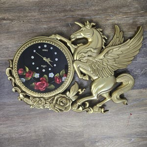 May include: Gold-tone wall clock featuring a winged unicorn design with a black clock face and red rose accents. The clock face reads "Linox Quartz".