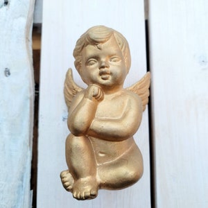 Puede incluir: Una figurita de querubín dorada con alas, sentada con los brazos cruzados y una expresión pensativa en su rostro.