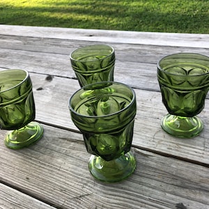 May include: Four green glass drinking glasses with a textured design and a short stem. The glasses are sitting on a wooden surface.