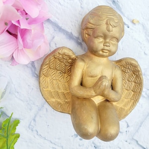 May include: A gold-colored cherub figurine with wings, kneeling with hands clasped in prayer. The figurine is made of ceramic or plaster and has a detailed, realistic design.
