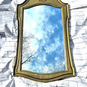 May include: A large, ornate gold-painted mirror with a decorative top and a beveled edge. The mirror reflects a blue sky with white clouds and tree branches.