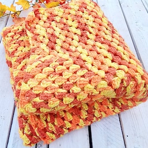 May include: A crocheted blanket in shades of orange, yellow, and peach. The blanket is folded in half and has a textured, waffle-like pattern.
