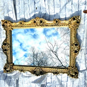 May include: A large, ornate gold mirror with a heavily carved frame. The mirror reflects a blue sky with white clouds and tree branches.