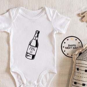 Puede incluir: Un body blanco para bebé con una ilustración negra de una botella de cerveza con el texto "IPA Bit" en ella.