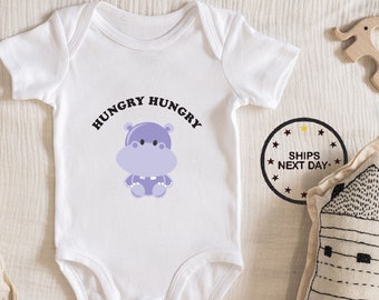 May include: White baby onesie with a purple hippopotamus illustration and the text "Hungry Hungry" printed on the front.
