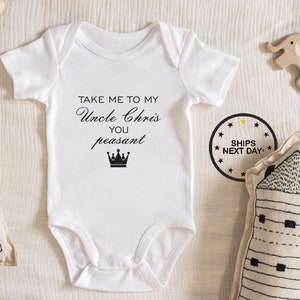 May include: White baby onesie with black text that reads "Take me to my Uncle Chris, you peasant" with a crown graphic.