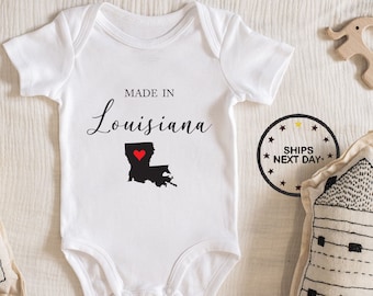 May include: White baby onesie with the text "Made in Louisiana" and a black outline of the state of Louisiana with a red heart in the center.