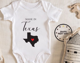 May include: White baby onesie with black text that reads "Made in Texas" and a black outline of the state of Texas with a red heart in the center.