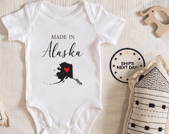 May include: White baby onesie with black text that reads "Made in Alaska" and a black outline of the state of Alaska with a red heart in the center.