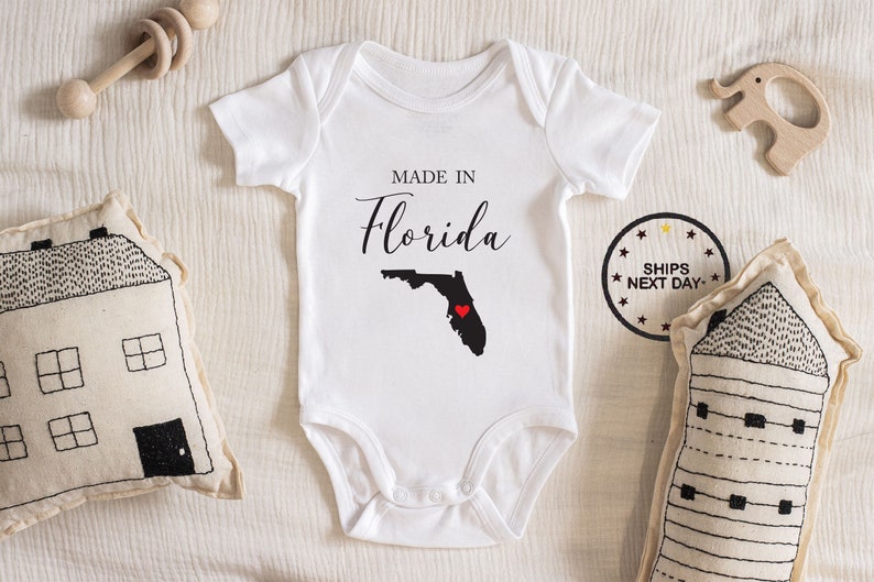 May include: White baby onesie with the text "Made in Florida" and an outline of the state of Florida with a red heart in the center.