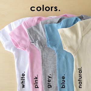 May include: Five baby onesies in different colors: white, pink, grey, blue, and natural. The onesies are laid out in a row, with the white onesie on the left and the natural onesie on the right. The text "colors." is written above the onesies.