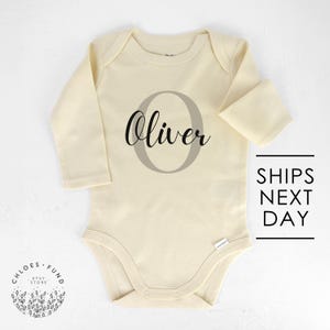 May include: A cream-colored long-sleeved baby onesie with the name "Oliver" written in black cursive inside a large gray circle.